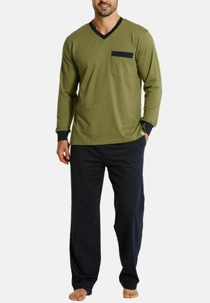 Mann trägt ein olivgrünes Langarmhemd mit schwarzem Besatz und Brusttasche, kombiniert mit lockeren schwarzen Hosen, steht barfuß.