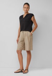 Schwarzes Kurzarmoberteil mit V-Ausschnitt und Details an den Schultern, kombiniert mit beigen, gefalteten Shorts, abgerundet mit schwarzen Sandalen.