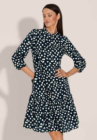 Navyblaues Kleid mit weißen Polka-Dots, Knopfverschluss, Kragen, Dreiviertelärmel und einem schichten Rüsche-Saum. Baumwollstofftextur.