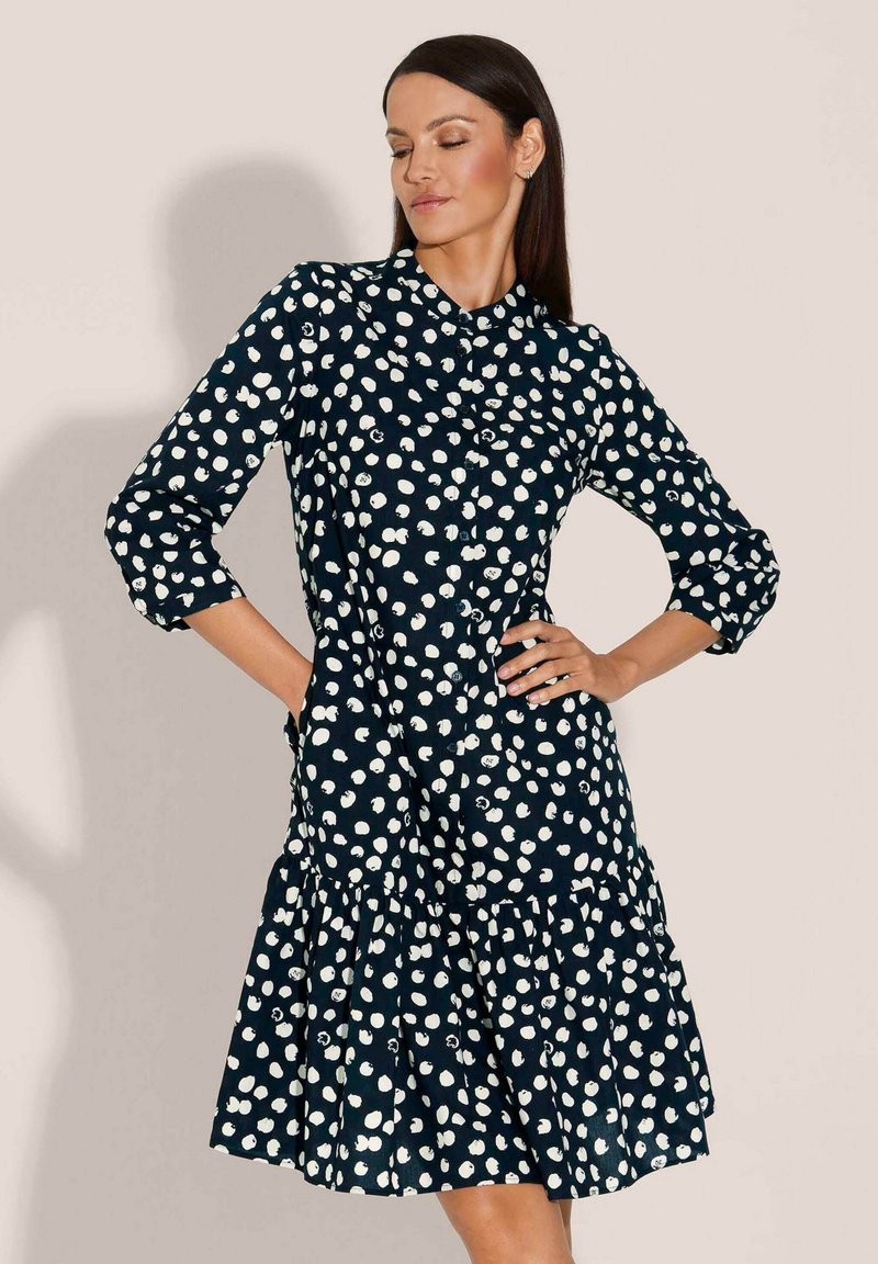 Navyblaues Kleid mit weißen Polka-Dots, Knopfverschluss, Kragen, Dreiviertelärmel und einem schichten Rüsche-Saum. Baumwollstofftextur.