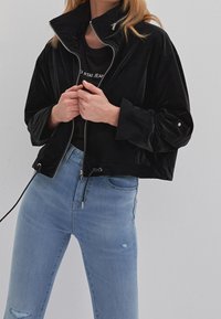 Kvinna i en svart kort zip-up jacka över en svart t-shirt och ljusblå jeans med hög midja och mindre slitningar.