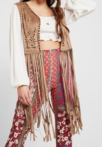 Braune Wildlederweste mit Fransen und lasergeschnittenen Mustern, getragen über einem weißen Oberteil und bunten gemusterten Hosen mit floralen und geometrischen Designs.