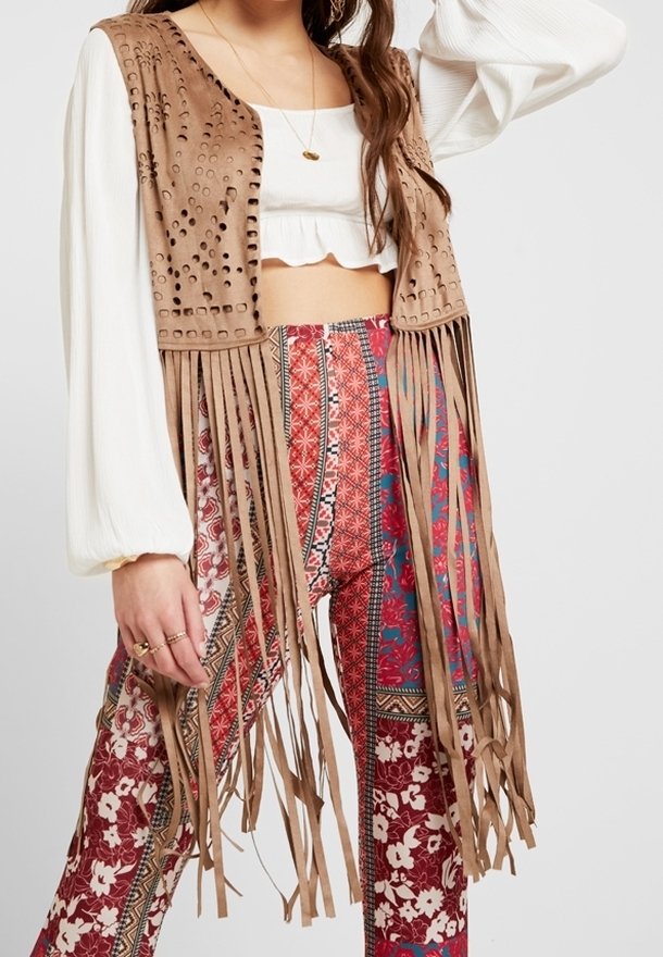 Braune Wildlederweste mit Fransen und lasergeschnittenen Mustern, getragen über einem weißen Oberteil und bunten gemusterten Hosen mit floralen und geometrischen Designs.