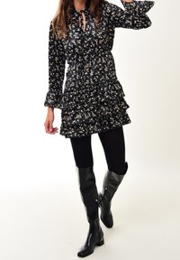 Schwarzes Blumenkleid mit gerafftem Saum, langen Ärmeln und Bindekragen. Getragen mit schwarzen Leggings und hohen schwarzen Lederstiefeln mit Absatz.
