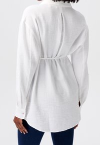 Chemise blanche texturée avec une coupe décontractée, des manches longues et un lien à la taille, dotée d'un col simple et de poignets boutonnés.