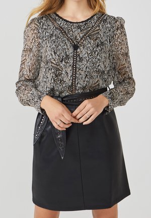 Mujer con blusa de manga larga transparente con estampado y falda de cuero negro con cinturón de tachuelas, manos entrelazadas a la altura de la cintura.