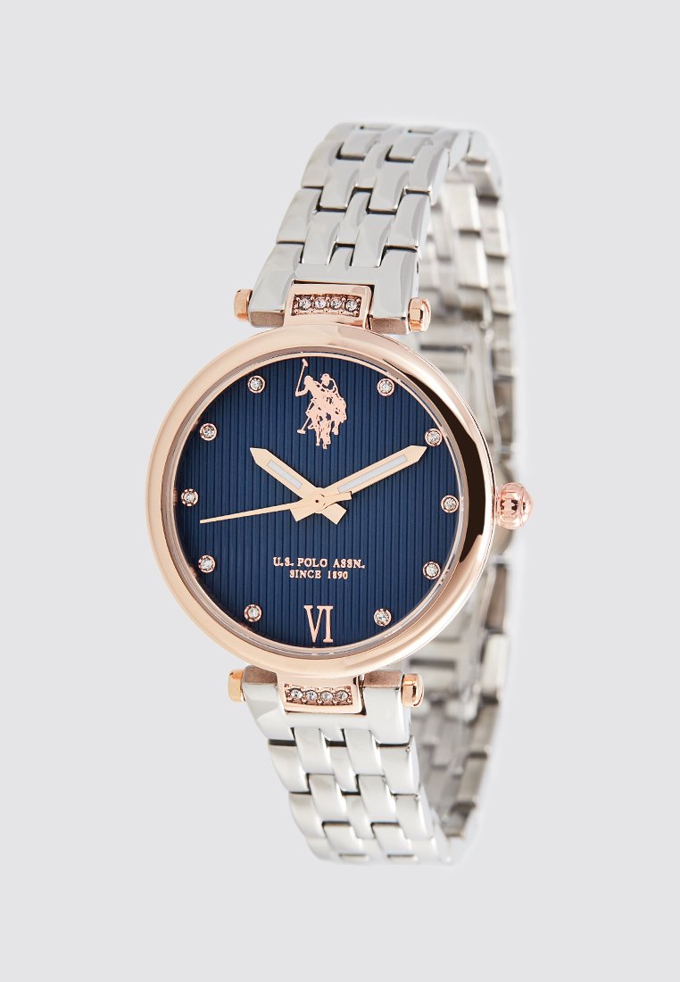 Reloj redondo con una esfera texturizada azul marino, acentos en oro rosado, marcadores de cristal y una correa metálica plateada. Presenta el logotipo de U.S. Polo Assn.