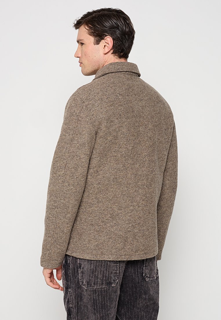 Braune Wollmischungjacke mit Kragen, die eine strukturierte Oberfläche und Nähte aufweist. Tragen über schwarzen vertikal gestreiften Hosen.