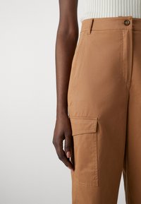 Hnědé cargo kalhoty s hladkým povrchem, obsahující boční kapsu, vysoký pas a jedno zapínání na knoflík. Materiál z bavlněné směsi.