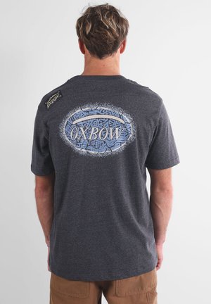 T-shirt gris à manches courtes avec un logo bleu et des motifs tribaux au dos, avec une petite étiquette de marque sur l'épaule gauche.