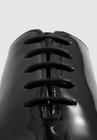 Scarpe in pelle verniciata nera con punta arrotondata, caratterizzate da sette stringhe nere attraverso gli occhielli, superficie liscia e lucida, e dettagli minimi.