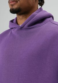Sweat à capuche violet en tissu doux et texturé. Dispose d'une large capuche doublée et d'une coupe décontractée avec des détails de couture visibles.