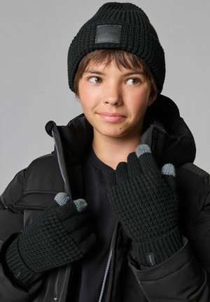 Schwarze Strickmütze mit einem Lederpatch und passenden fingerlosen Handschuhen mit grauen Spitzen. Die Handschuhe haben ein strukturiertes, geripptes Muster.