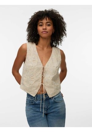 Kvinde med krøllet hår iført en ærmeløs beige broderet vest, bundet foran, og blå cowboybukser, stående med hænderne på ryggen.