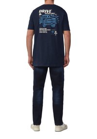 Marineblauwe shirt met grafische print op de rug, met een vrachtwagen en tekst. Gecombineerd met donkere spijkerbroeken en lichtgekleurde sneakers.