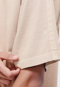 Beige short-sleeved shirt with a relaxed fit, made of soft fabric; features side stitching and "ALL SZN" embroidery near the hem.