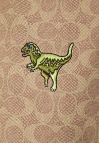 Patch dinosaure vert brodé avec des dents acérées sur un fond en tissu texturé présentant des motifs circulaires beiges répétitifs.
