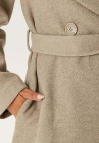 Helle beigefarbene Wollmischungjacke mit weicher Textur, ausgestattet mit einem taillierten Gürtel, großen Knopfverschluss und Seitentaschen.