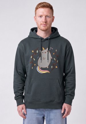 Homme portant un sweat à capuche foncé avec une licorne-chat de dessin animé entourée de feuilles et de branches, associé à un jean bleu clair.