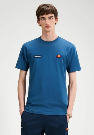 Jonge man die een blauw ellesse T-shirt en marineblauwe broek draagt, met zijn handen in de zakken, staand tegen een effen witte achtergrond.