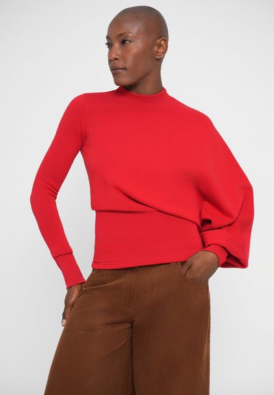 Camisa roja de manga larga con cuello alto y diseño de hombros drapeados, combinada con pantalones de pana marrón de pierna ancha. Textura suave, estilo ajustado.