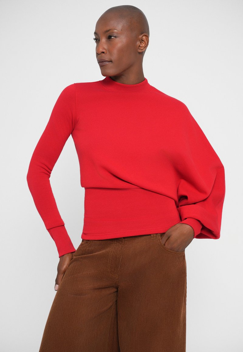 Red long-sleeve shirt with a high neck and draped shoulder design, paired with brown corduroy wide-leg pants. Smooth texture, fitted style.