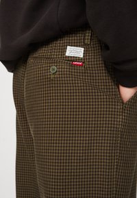 Bruine geruite broeken met een groene accentknop en een Levi's label op de achterzak. Gemaakt van duurzame stof met een soepele textuur.
