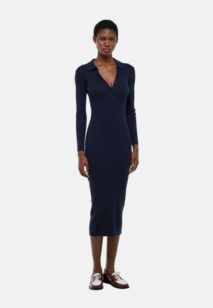 Femme portant une robe en maille côtelée bleu marine ajustée avec manches longues, col, encolure en V boutonnée, et des chaussures montantes bicolores à lacets.