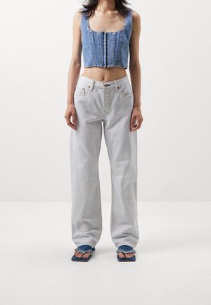 Person wearing a sleeveless cropped denim top, light-colored loose jeans, and blue square-toe sandals on a plain background.