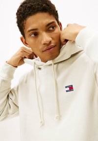 Sudadera color crema hecha de material suave, con capucha ajustable y un pequeño parche de logo azul, rojo y blanco en el pecho.