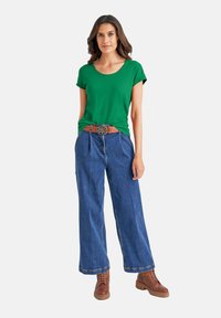Groene t-shirt met korte mouwen gecombineerd met wijde blauwe spijkerbroeken en bruine veterschoenen. Bruine riem met een decoratieve gesp.