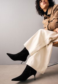 Black suede ankle boots with pointed toes and stiletto heels, paired with wide-leg cream trousers. Smooth texture and defined silhouette.