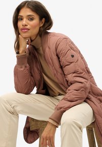 Femme assise sur un tabouret en bois, portant une veste matelassée rose poussiéreux, un pull col roulé beige et un pantalon beige, sur un fond blanc uni.