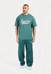 Turkos oversize t-shirt och matchande vida byxor med "Smilodox"-logotyp i vitt, gjorda av mjukt tyg med slät textur.