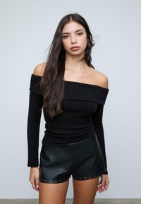 Mladá žena s dlhými tmavými vlasmi, oblečená v čiernom rebrovanom topu s odhalenými ramenami a čiernych kožených šortkách s kovovými cvokmi na lemu.