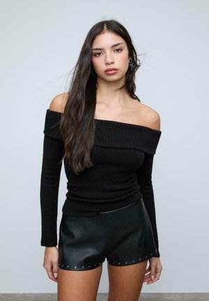 Junge Frau mit langen dunklen Haaren, die ein schwarz geriffeltes Top mit schulterfreier Optik und schwarze Leder-Shorts mit Metallnieten am Saum trägt.