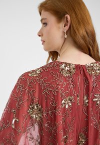 Abito rosso ricamato con motivi floreali e fogliari, adornato con paillettes. Presenta una chiusura con cerniera sul retro e una texture di tessuto trasparente.
