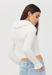 Biały sweter z prążkowanej tkaniny z kołnierzem z sztucznego futra, krótki fason, z długimi rękawami, w połączeniu z jasnoniebieskimi dżinsami.