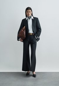 Must kohandatud ülikond laia lõikega pükste ja valge särgiga, mustade kontsadega. Välimuse lõpetavad pruun kott ja vöö. Sile kangas, struktureeritud disain.