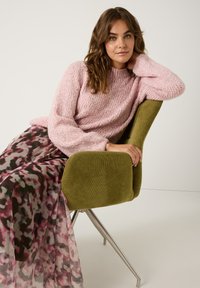 Rosa gestrickter Pullover mit hohem Kragen, kombiniert mit einem durchsichtigen, gemusterten Kleid in Rosa-, Lila- und Grüntönen. Sitzt in einem grünen Stuhl.