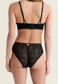 Dos d'une femme portant une culotte taille haute en dentelle noire à motif floral et un soutien-gorge noir assorti avec bretelles réglables, fond beige.