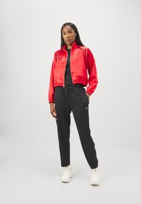 Sarkans satīna jaciņa ar rāvējslēdzēju un pieguļošu vidukli, melns T-krekls un melni treniņbikses ar sānu pogām, apvienoti ar baltām sporta kedām.