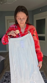 White skirt made of lightweight fabric, featuring gathered details and a waistband. Hangs on a hanger, with a visible tag attached.