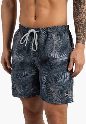 Homme portant un short de bain foncé avec un motif de feuille de palmier blanc, un cordon blanc et un tatouage tribal sur l'avant-bras droit.