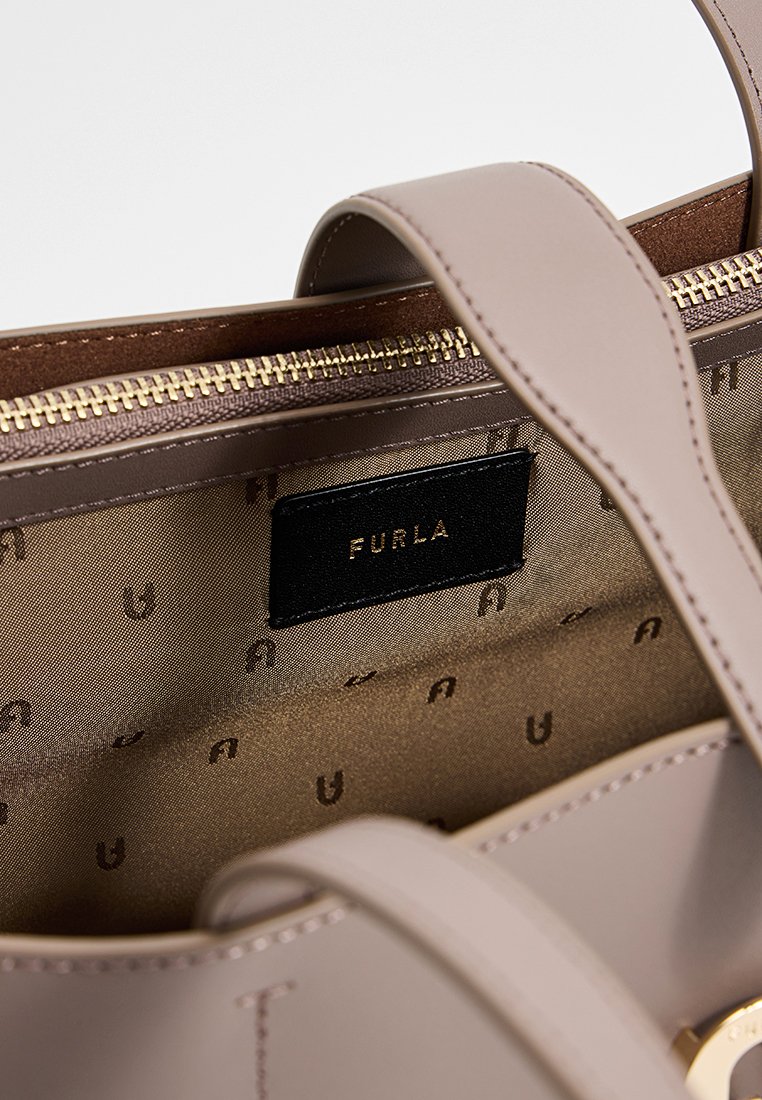 Hellebeige Lederhandtasche mit goldfarbenem Reißverschluss, ausgestattet mit einem braunen Innenfutter, das mit einem Logomuster geprägt ist. Schwarzes Markenlabel innen sichtbar.