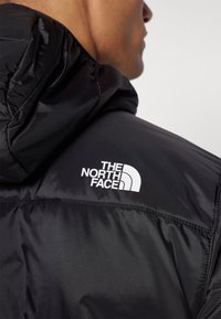 Svart hettejakke med teksturert finish, synlige sømmer og den hvite logoen til "The North Face" på venstre skulder.