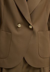 Blazer marrón de doble botonadura hecho de tela suave, con botones dorados en forma de león, dos grandes bolsillos frontales y solapas afiladas.