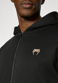 Μαύρη φούτερ ζακέτα με φερμουάρ και απαλό ύφασμα, με μικρό λογότυπο φιδιού σε καφέ απόχρωση στην αριστερή πλευρά του στήθους. Είναι ορατές λεπτομέρειες του φερμουάρ και της κουκούλας.