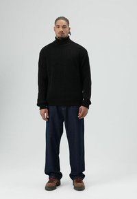 Man die rechtop staat en naar voren kijkt, gekleed in een zwarte rolkraagtrui, losse donkere spijkerbroek en bruine wandelschoenen met rode veters, tegen een effen lichte achtergrond.