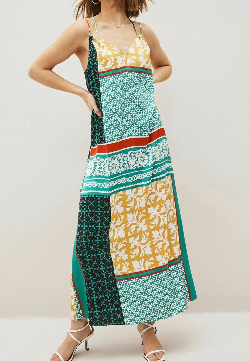 Femme portant une robe maxi sans manches multicolore avec des motifs floraux et géométriques, debout les mains sur les hanches et des talons blancs à brides.